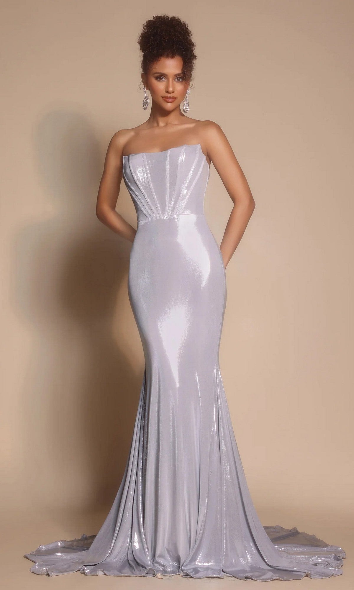 A woman wears the Portia and Scarlett XO097 long formal dress—a strapless, silver metallic gown with a fitted silhouette and floor-length train—standing against a plain beige background.